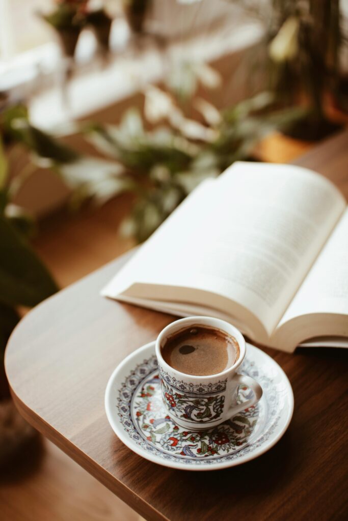 A beautifully ornate cup of Turkish coffee sits next to an open book on a wooden table, surrounded by indoor plants.