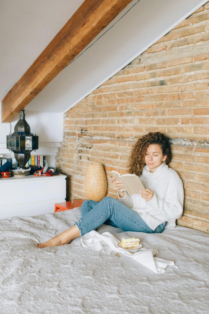 Woman enjoying leisure time reading book in a cozy, rustic bedroom setting with warm ambiance.